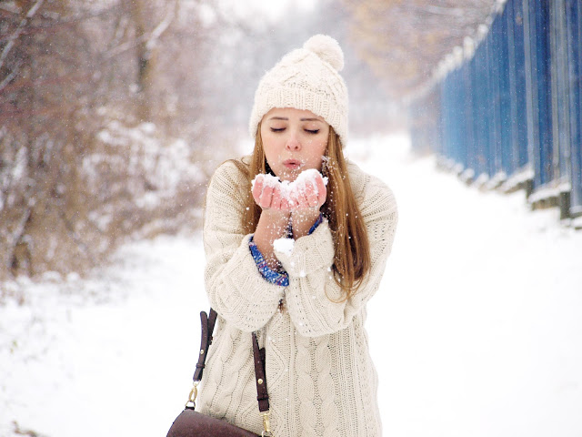 Девушка в белом свитере и шапке сдувает с рук снег
