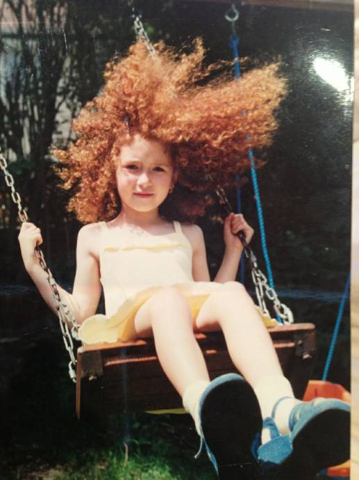Девочка с кудрявыми рыжими волосами на качелях